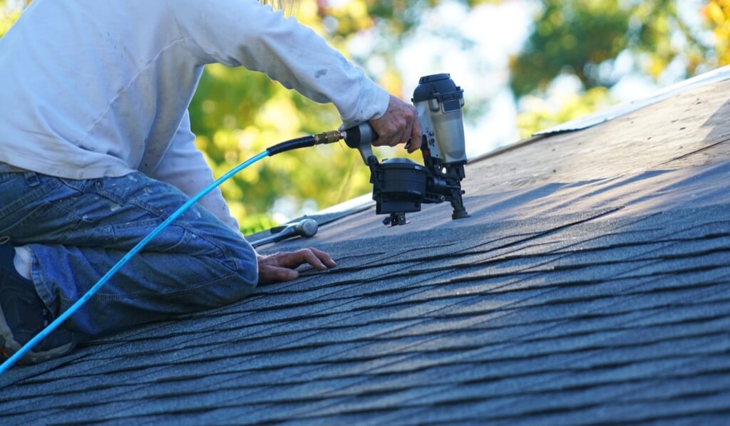 A roofing worker is using a nail gun to nail shingles to a roof deck.